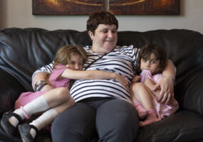 Charmaine, a Caucasian woman with short dark brown hair, is sitting on a sofa being cuddled by her daughters. Charmaine is smiling and the girls are looking shyly at the camera.