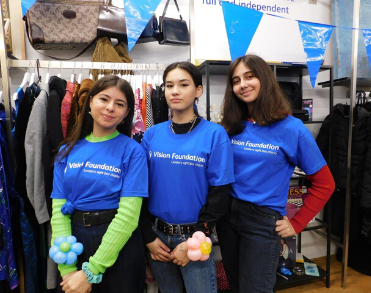 Three young women pose in a charity shop wearing blue Vision Foundation t-shirts