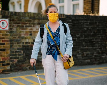 A lady, Odette, walking down the street with a white cane. Odette wears an orange face mask due to the Covid-19 pandemic.