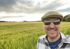 Chris standing in front of a field of wheat. The corn is turning golden and it's partially cloudy day. You can see some bright sunshine behind grey clouds. Chris is wearing a flat cap, sunglasses and a checked shirt over a blue tshirt.