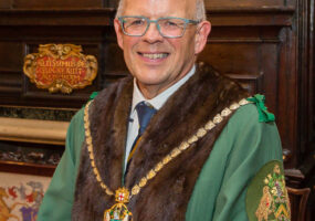 Ian Davies a white man with white hair and glasses. He is wearing the robes and livery of the Company