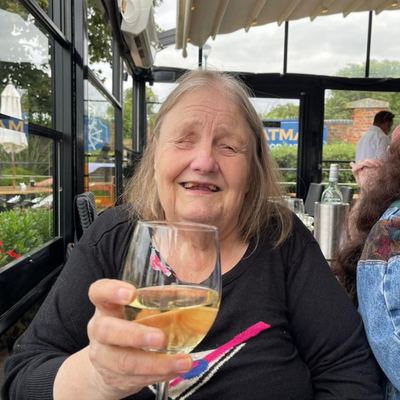 Carol an older woman with grey hair smiles at the camera holding a glass of wine. She's wearing a black shirt and sitting at a table in a restaurant
