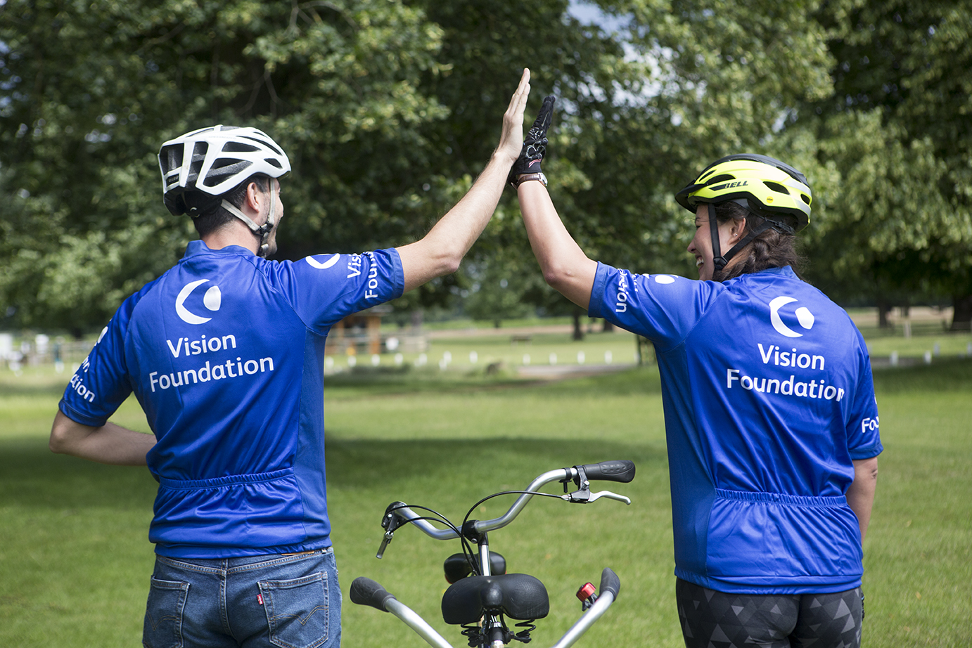 Two people wearing cycling helmets and blue Vision Foundation branded cycling jerseys are facing away from the camera and high fiving each other. They are outside in a park and there is a tandem bicycle between them.