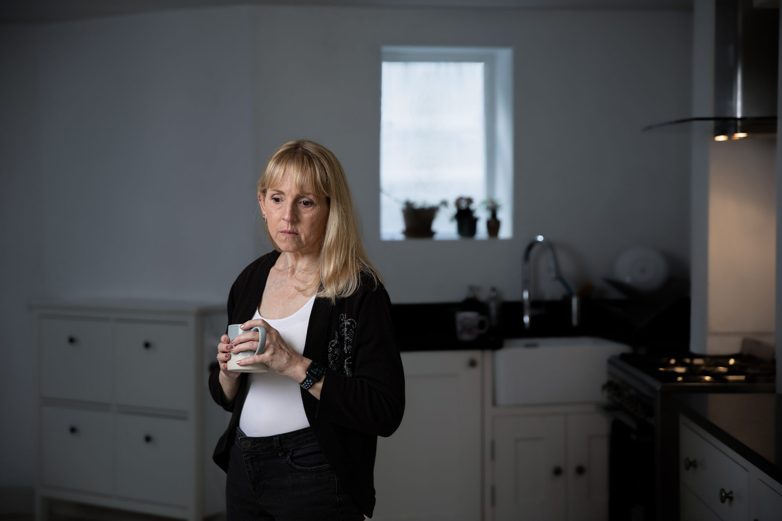 A blond woman stands in her kitchen holding a mug of tea. She is looking down at the floor.