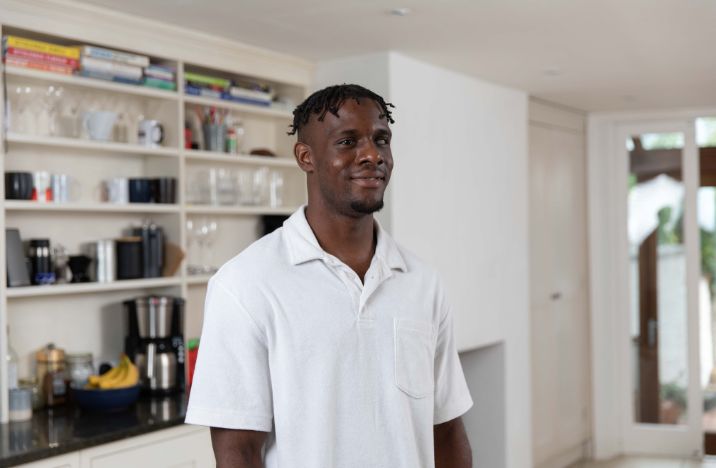 A young black man stands in a kitchen. He stands proudly with a small smile on his face and is wearing a white polo neck shirt