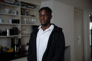 Fikayo, a young black man stands in a living room looking down at the floor. He's wearing a white shirt and black hoodie