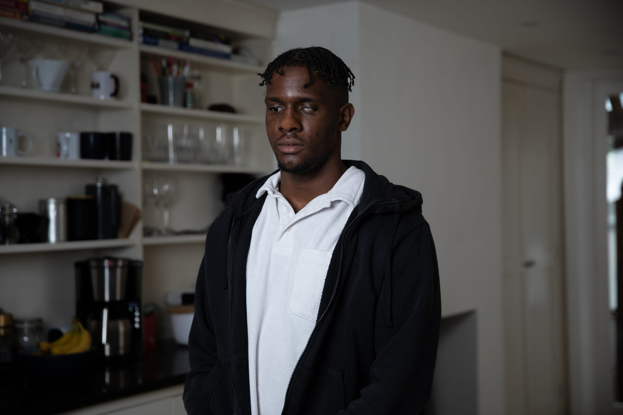 Fikayo, a young black man stands in a living room looking down at the floor. He's wearing a white shirt and black hoodie