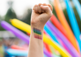 A raised fist of a white person against a background of rainbow-coloured tube-shaped balloons. On their wrist, they have the colours of the pride flag painted on.