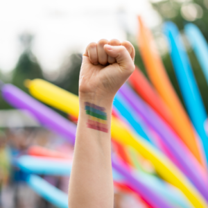 A raised fist of a white person against a background of rainbow-coloured tube-shaped balloons. On their wrist, they have the colours of the pride flag painted on.