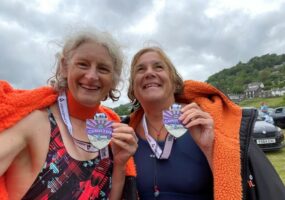Monica and her friend smiling for a post swim selfie. Wrapped in fuzzy coats, holding their medals and smiling at the camera.