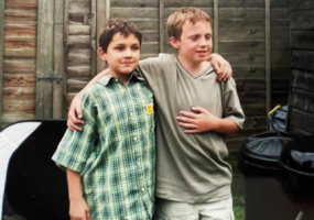 A young Joe K and Joe P standing arm-in-arm for a photo in a fenced garden. Joe K has short, dark brown hair and wears a green, blue and cream coloured plaid button-up shirt. Joe P has short, auburn hair and is wearing a khaki-brown v-neck t-shirt.