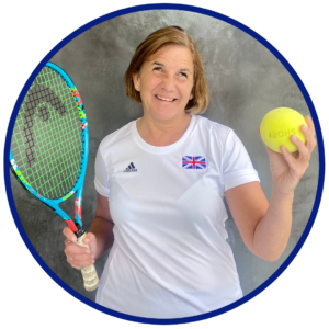 Monica Smith posing against a grey background. She is wearing a white Team GB tennis top with the flag embroidered on her chest. She is smiling casually whilst holding her tennis ball and racket up for the camera.