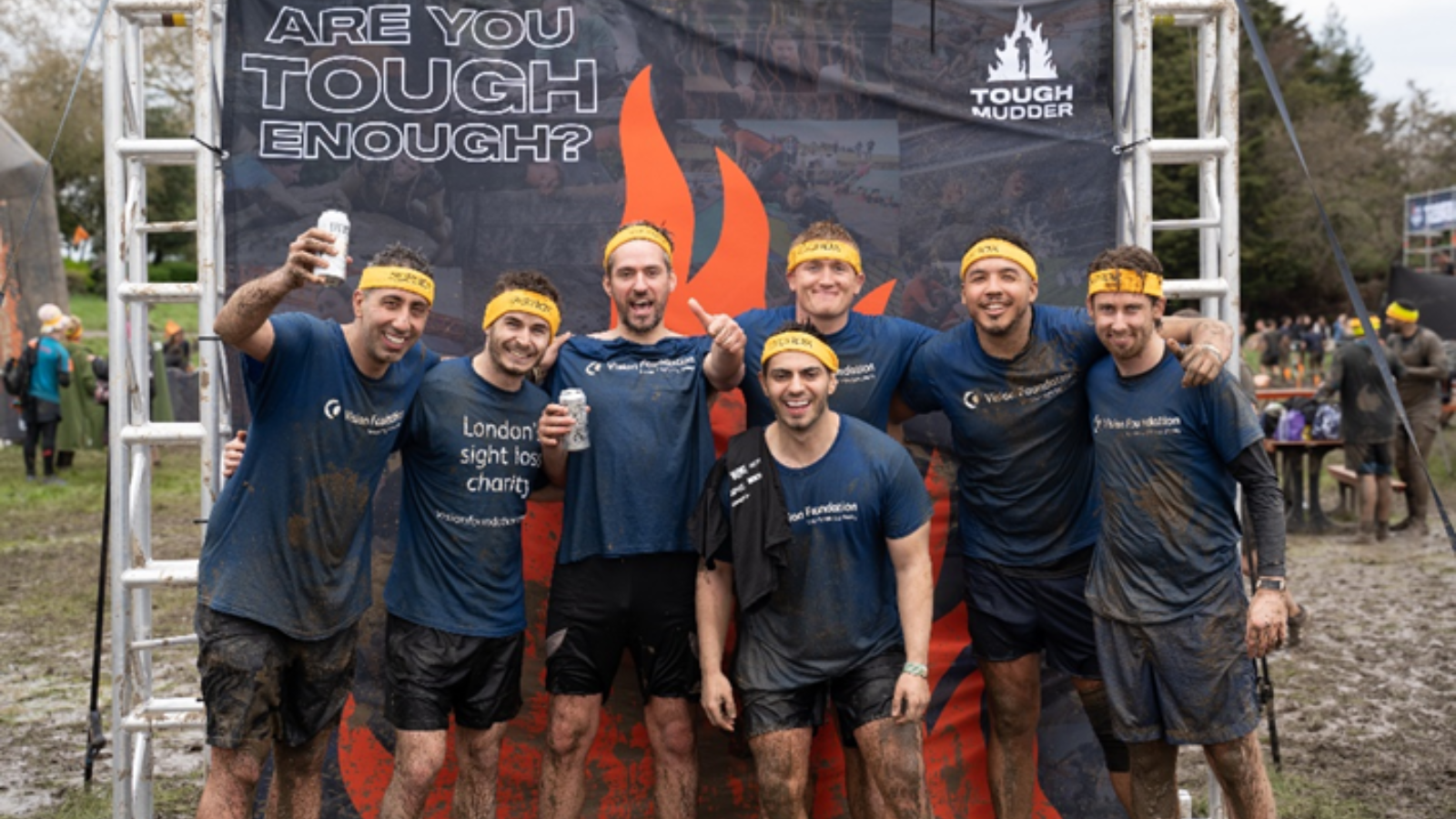 Seven men in vision foundation shirts and yellow bandanas standing in front of an 'are you tough enough?' Tough Mudder promotional banner. They're all grinning and covered in mud.