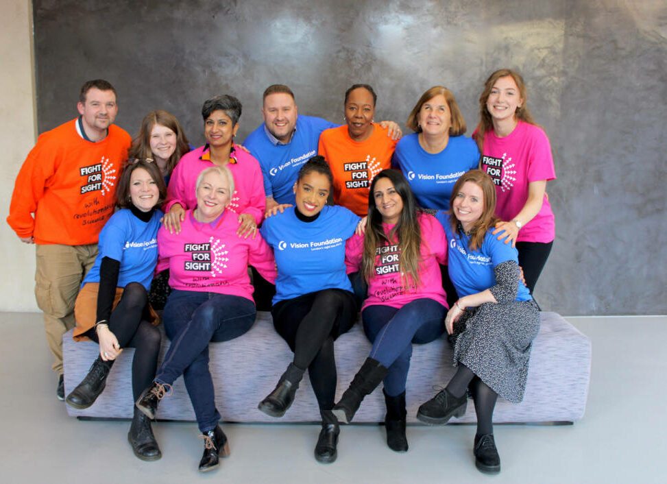 Two rows of colleagues posing closely for a smiley photo in an office foyer. The colleagues are wearing a mix of colourful T-shirts, including blue VF tees and pink and orange FFS tees and sweatshirts.
