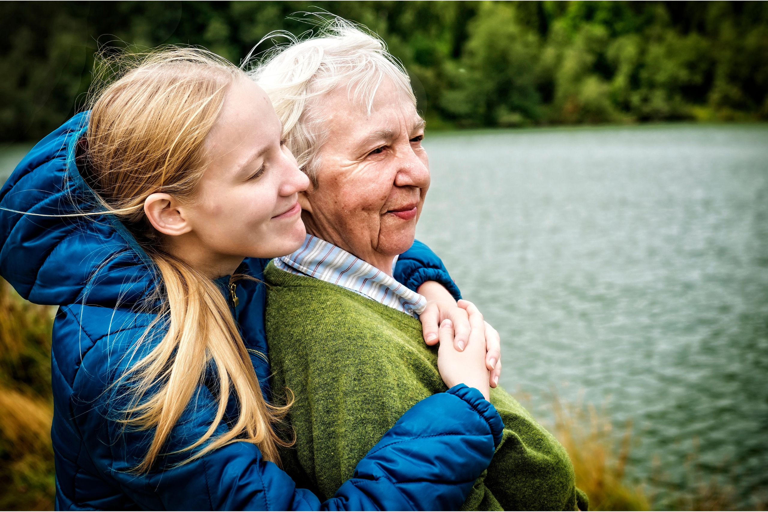 A teenager hugging an older lady from behind as they look out at a lake.