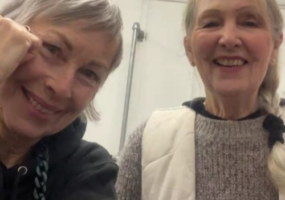 Two women smile into the camera, both have grey hair. One woman wears a black top and the second woman wears a grey jumper.